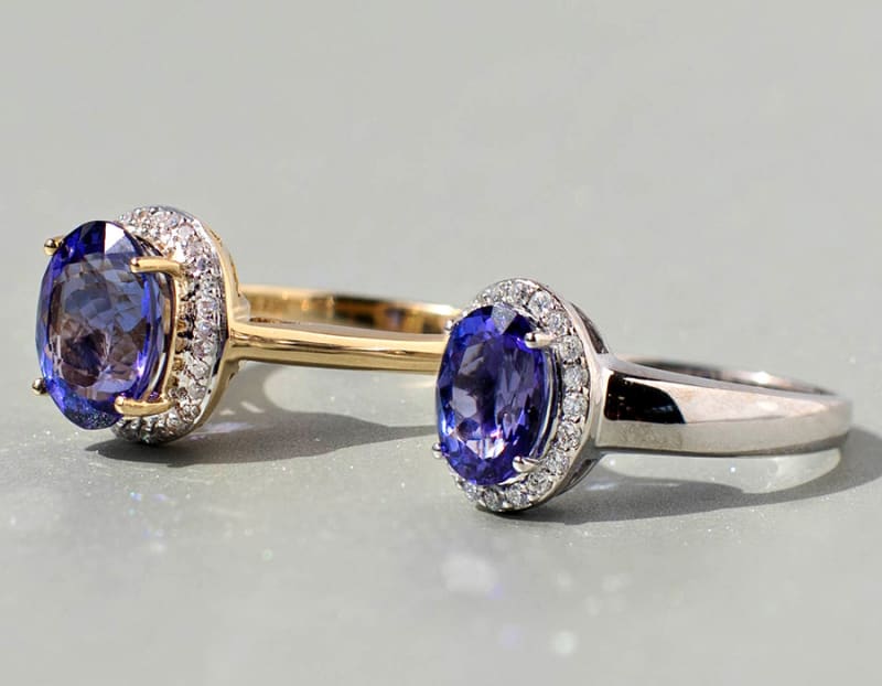 This photo is a close up of two tanzanite rings. The first ring is an oval tanzanite ring with round white diamonds around it on a yellow gold setting. The second ring is an oval tanzanite ring with round white diamonds around it on a white gold setting. 