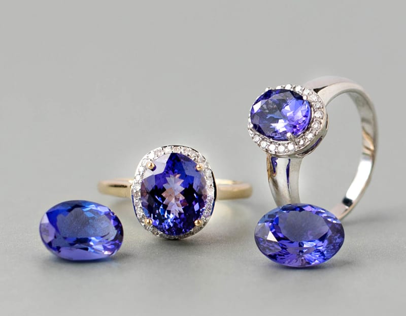 This picture is a close up of two tanzanite rings and two tanzanite gemstones. The first ring is an oval tanzanite stone with round, white diamonds around it on a white gold setting, the second is an oval tanzanite ring with round white diamonds around it on a yellow gold setting. The first gemstone is signature tanzanite stone and the second is an heirloom tanzanite stone. 