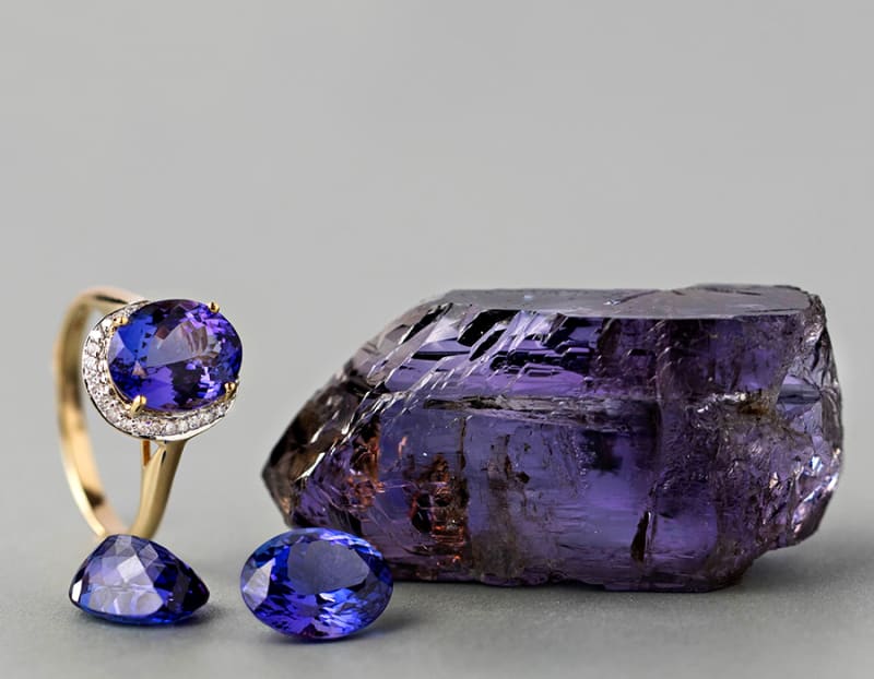 A gold tanzanite ring with two loose tanzanite stones next to a dark purple blue tanzanite stone. 