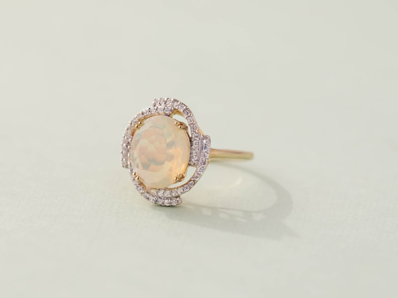 Close-up of a multi-color Ethiopian opal yellow hold ring. The main stone is circular and has small arms adorned with small diamonds spiraling off of it.  