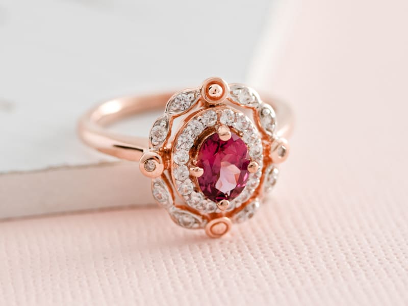 Close-up of a pink garnet ring set in rose gold, with the main stone surrounded by a plethora of small white accent stones. The ring is shot on a soft pink and white background. 