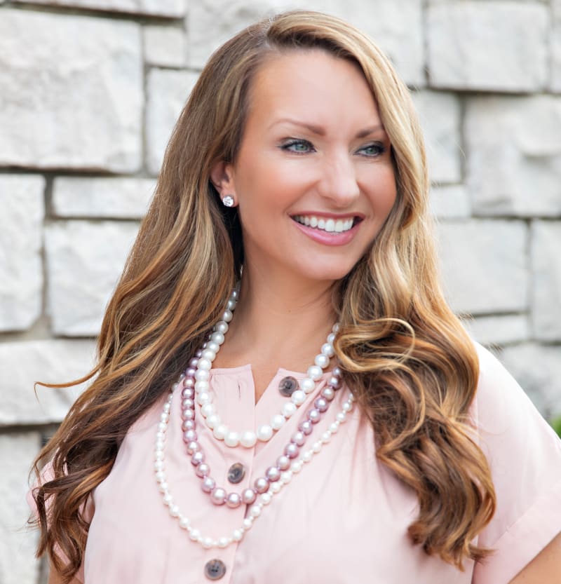 A brown haired woman wearing a pink shirt and three pearl strand necklaces, two white pearls and one with pink pearls.  