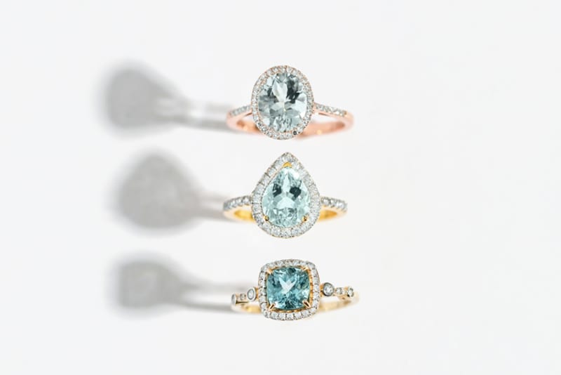 A line of three aquamarine rings stacked on top of each other, photographed on a white background. From top to bottom: circular cut, square cut and teardrop cut gemstones. 