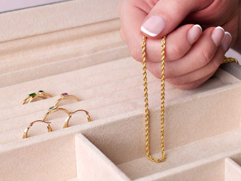 A hand holds a gold rope chain with gold earrings and rings behind it. 