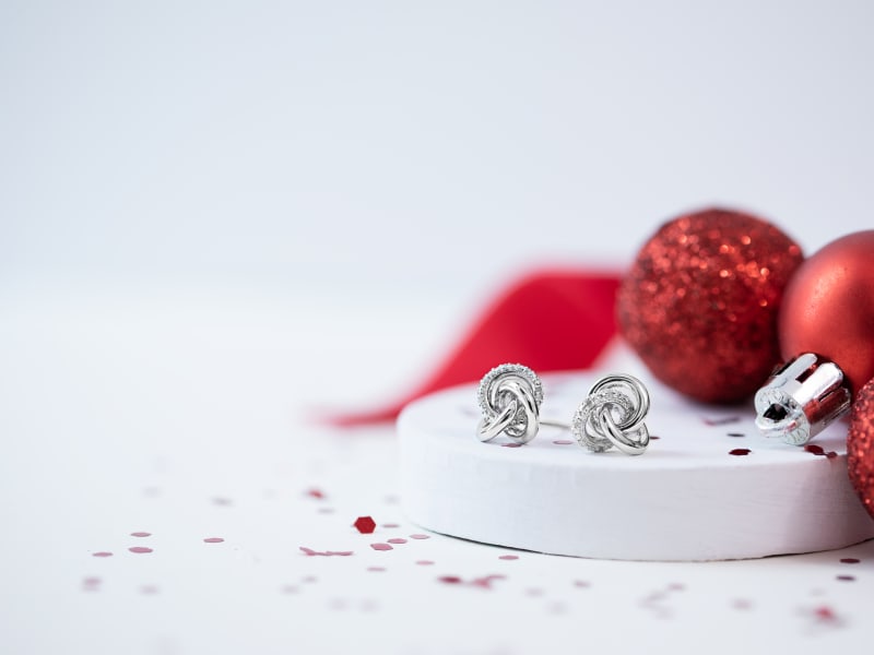 A pair of silver knot-shaped stud earrings with red ornaments and ribbon in the background 