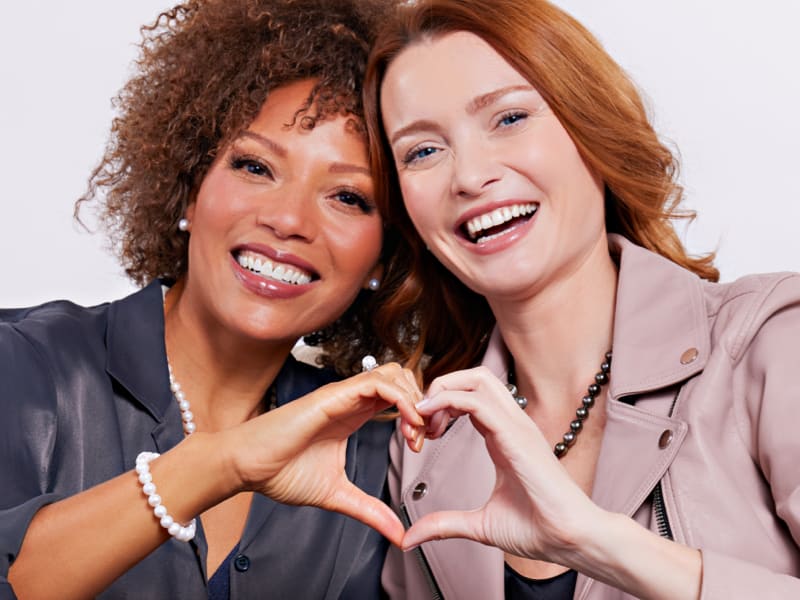 Two female friends sitting on a couch making a heart symbol with their hands 