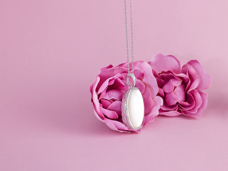 A silver and white locket hanging in front of a pink flower 